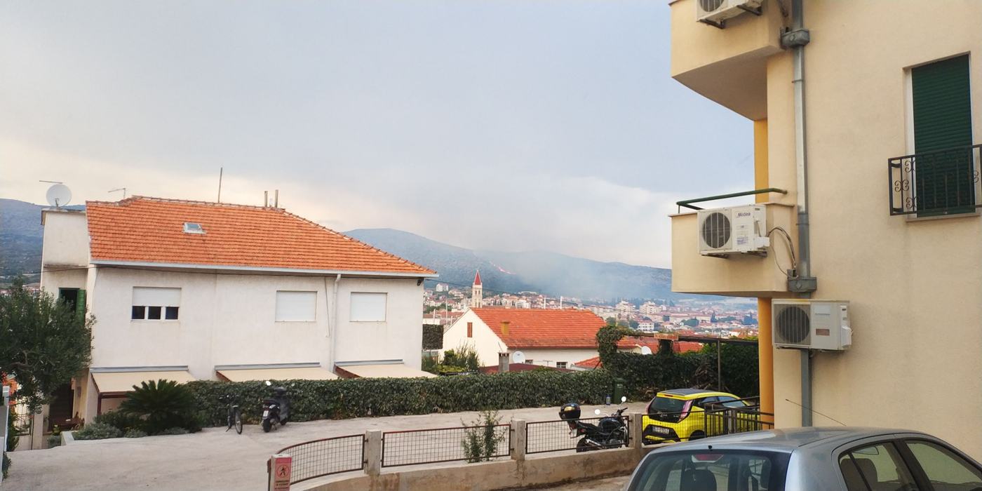 Rauch und eine Feuerlinie am Hang über einer Küstenstadt, gesehen aus einer Wohnstraße zwischen Häusern